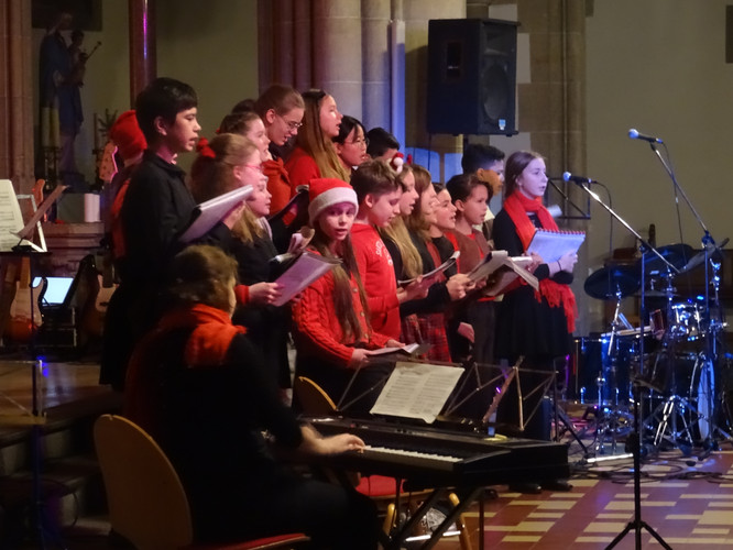 KI generiert: Das Bild zeigt einen Chor von jungen Sängerinnen und Sängern, die während einer Aufführung in einer Kirche Weihnachtslieder vortragen. Sie tragen festliche Kleidung, darunter einige mit weihnachtlichen Accessoires wie Nikolausmützen.