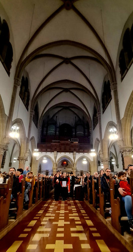 KI generiert: Das Bild zeigt das Innere einer Kirche mit hohen Gewölbedecken und Reihen von Bänken, die mit Menschen gefüllt sind. Im Mittelgang schreiten zwei Personen in schwarzen Talaren feierlich nach vorne.