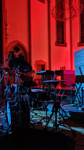 KI generiert: Das Bild zeigt ein Schlagzeug und eine Keyboard-Ausrüstung, die in einem rot beleuchteten Innenraum, möglicherweise einer Kirche, aufgestellt sind. Im Hintergrund sind hohe Fenster und Kerzenleuchter erkennbar.