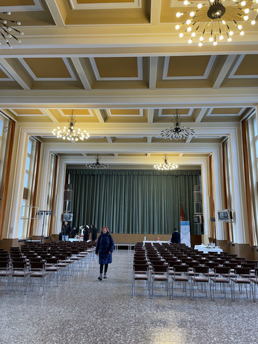 KI generiert: Das Bild zeigt einen großen, eleganten Saal mit einer Bühne im Hintergrund und mehreren Reihen von leeren Stühlen, die auf eine Veranstaltung vorbereitet scheinen. Eine Person im Vordergrund geht auf die Kamera zu.