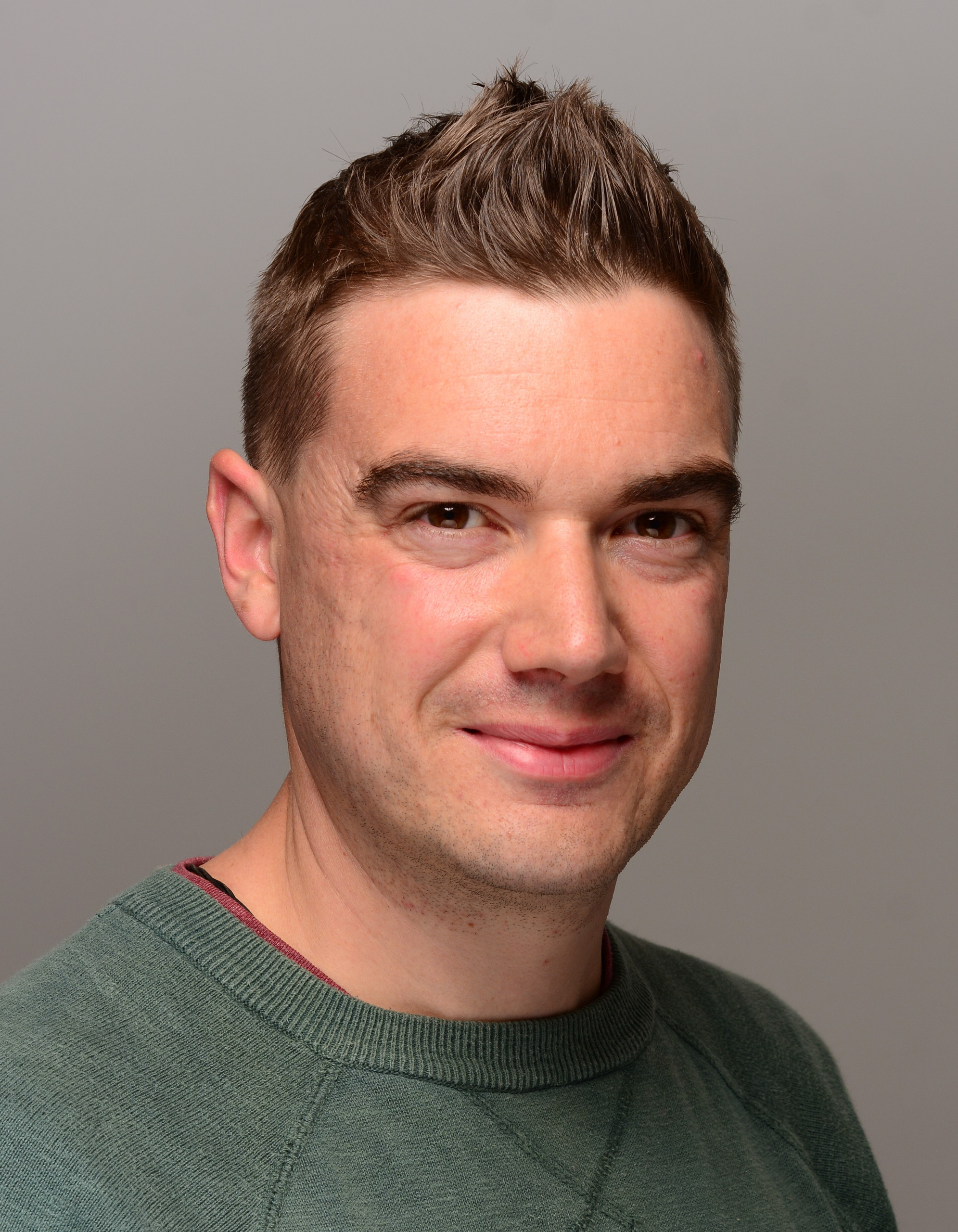 KI generiert: Das Bild zeigt einen Mann in einem grünen Pullover vor einem neutralen, grauen Hintergrund. Der Mann hat kurzes, nach oben gestyltes Haar und lächelt leicht.