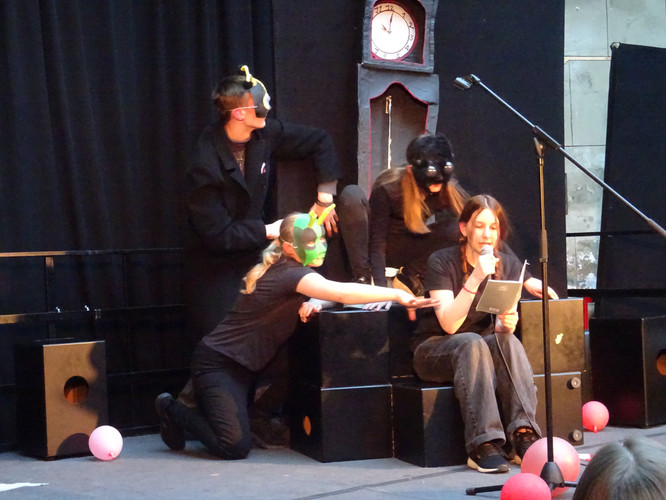 KI generiert: Auf dem Bild sind vier Personen zu sehen, die maskiert auf einer Bühne in einer Theaterdarbietung agieren. Eine Person sitzt und liest von einem Blatt, während die anderen drei in verschiedenen Posen um sie herum arrangiert sind.