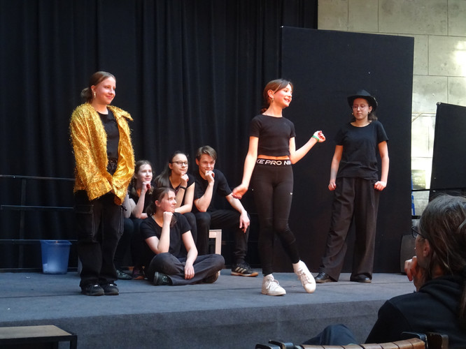 KI generiert: Das Bild zeigt eine Gruppe von Menschen auf einer Bühne, die offenbar an einer Theatervorstellung teilnehmen. Eine Person im Vordergrund scheint zu lächeln und zu gestikulieren, während andere zuschauen.