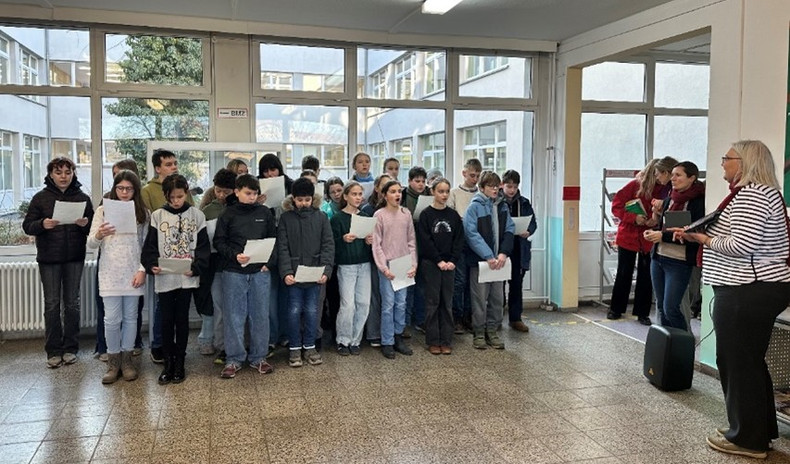 KI generiert: Eine Gruppe von Schülerinnen und Schülern steht in einem Innenbereich und hält Blätter in den Händen, während sie singen oder präsentieren. Auf der rechten Seite stehen zwei Erwachsene, von denen eine Person ein Mikrofon hält.