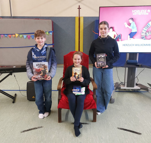 KI generiert: Auf dem Bild sind drei Kinder zu sehen, die jeweils ein Buch halten. Sie stehen und sitzen in einem Raum mit Dekorationen und einem Bildschirm, der auf einen Vorlesewettbewerb hinweist.
