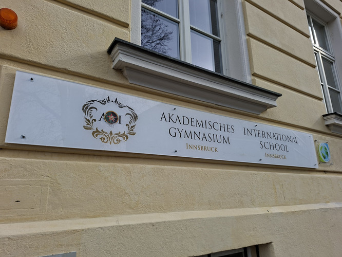KI generiert: Das Bild zeigt ein Schild an einer Gebäudeaußenwand mit der Aufschrift "Akademisches Gymnasium Innsbruck International School". Es handelt sich offenbar um den Eingang oder Teil der Fassade einer Bildungseinrichtung.