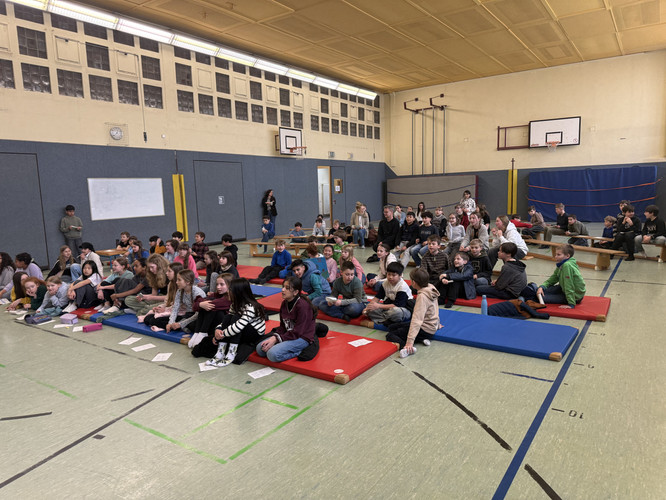 KI generiert: Das Bild zeigt eine Gruppe von Menschen, hauptsächlich Kinder, die auf Matten und Bänken in einer Turnhalle sitzen. Es wirkt wie eine Versammlung oder Schulveranstaltung.