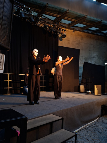 KI generiert: Das Bild zeigt eine Theaterbühne, auf der zwei Personen in dunkler Kleidung und weißen Masken eine Performance aufführen. Sie stehen mit ausgestreckten Armen in einer dramatischen Pose auf der Bühne.