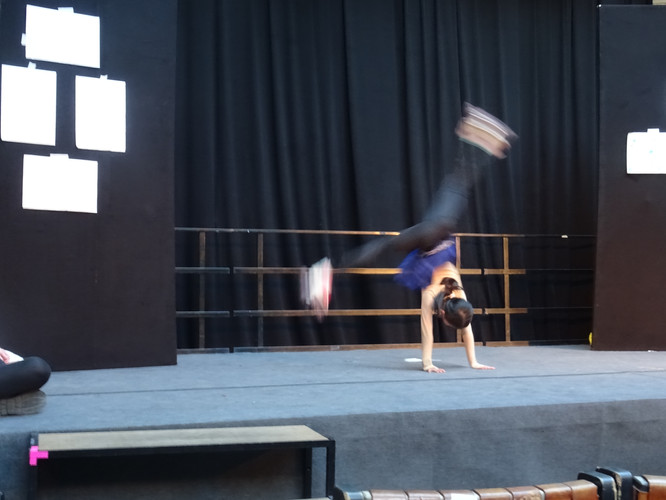 KI generiert: Das Bild zeigt eine Person, die auf einer Bühne einen Handstand oder akrobatischen Trick ausführt. Die Bühne ist von dunklen Vorhängen umgeben und im Hintergrund sind einige Poster zu sehen.