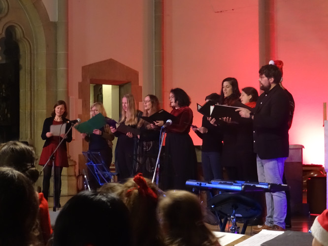 KI generiert: Das Bild zeigt eine Gruppe von Menschen, die auf einer Bühne in einem Innenraum singen. Sie halten Notenblätter in den Händen und es herrscht eine festliche Atmosphäre.
