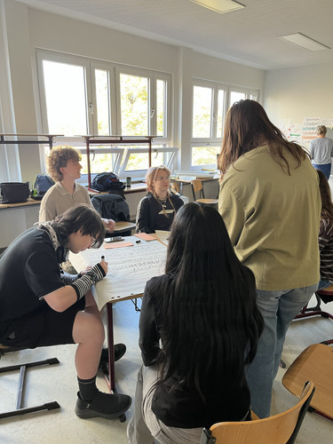 KI generiert: Eine Gruppe von Personen sitzt in einem hellen Raum um einen Tisch und arbeitet gemeinsam an einem großen Blatt Papier. Sie scheinen sich in einem schulischen oder akademischen Umfeld zu befinden.