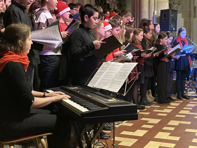 KI generiert: Das Bild zeigt einen Chor, der in einer festlichen Umgebung singt, begleitet von einer Person am Keyboard. Die Sänger tragen teils weihnachtliche Kleidung, was auf eine saisonale Aufführung hinweist.