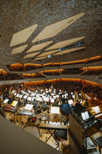 KI generiert: Das Bild zeigt ein Konzert in einem modernen, gut beleuchteten Konzertsaal, in dem ein großes Orchester vor einem gefüllten Publikum auftritt. Die Decke ist mit reflektierenden Flächen versehen, die einen beeindruckenden Lichteffekt erzeugen.