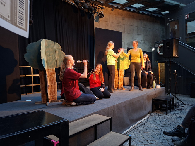 KI generiert: Das Bild zeigt eine Gruppe von Menschen auf einer kleinen Bühne, die offenbar ein Theaterstück oder eine Aufführung mit selbstgemachten Kulissen aufführen. Zwei Personen sitzen mit Mikrofonen, während die anderen interagieren, wobei im Hintergrund Bäume aus Pappe zu sehen sind.