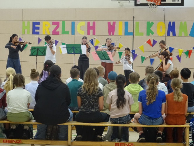 KI generiert: Das Bild zeigt eine Gruppe von Menschen, die auf einer Bühne mit Musikinstrumenten auftreten, während ein Publikum sitzt und zuschaut. Der Hintergrund ist mit einem "HERZLICH WILLKOMMEN"-Banner dekoriert.
