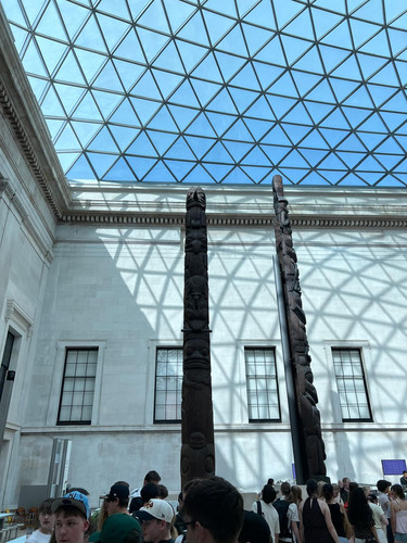 KI generiert: Das Bild zeigt zwei hohe Totempfähle in einem Gebäude mit einem beeindruckenden Glasdach, durch das Tageslicht hereinfällt. Im Vordergrund sind viele Menschen zu sehen, die sich die Ausstellung ansehen.