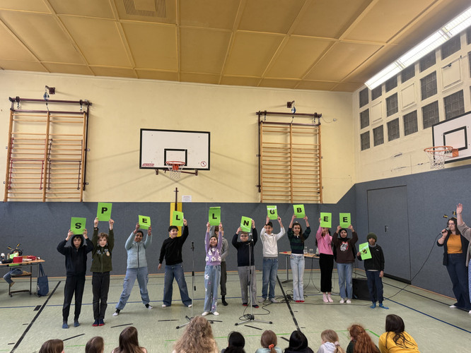 KI generiert: Eine Gruppe von Menschen steht in einer Turnhalle und hält Buchstaben hoch, die zusammen "SPELLING BEE" ergeben. Sie scheinen an einer Veranstaltung oder Präsentation vor einem Publikum teilzunehmen.