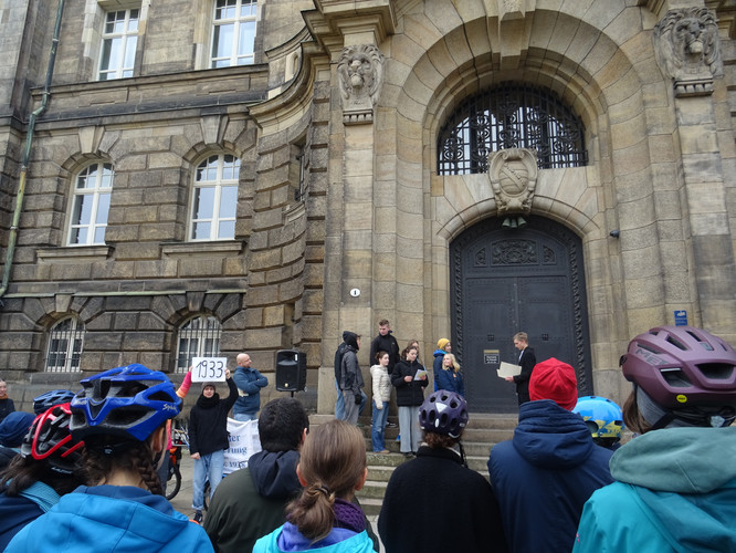 KI generiert: Auf dem Bild versammelt sich eine Gruppe von Menschen vor einem historischen Gebäude, wobei einige von ihnen Plakate und Schilder mit einer Jahreszahl hochhalten. Die Szene deutet auf eine Kundgebung oder Protestveranstaltung hin.