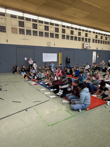 KI generiert: Das Bild zeigt eine Gruppe von Kindern, die in einer Turnhalle auf Matten sitzen und aufmerksam etwas beobachten oder zuhören. Im Hintergrund sind einige Erwachsene und Sportgeräte zu sehen.