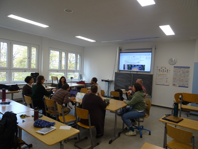 KI generiert: Das Bild zeigt eine Gruppe von Personen, die in einem Klassenzimmer um Tische herum sitzt und offenbar an einer Besprechung oder Schulung teilnimmt. Im Hintergrund ist eine große Anzeige oder Präsentation zu sehen.