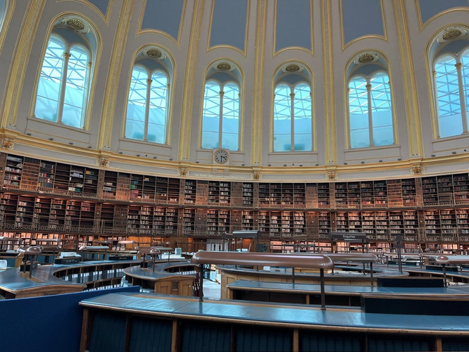 KI generiert: Das Bild zeigt einen großen, kreisförmigen Lesesaal mit hohen Decken und vielen Büchern in Regalen, die die Wände säumen. Zentral befindet sich ein Uhr, während die Architektur durch große Fenster, die viel Licht einlassen, geprägt ist.