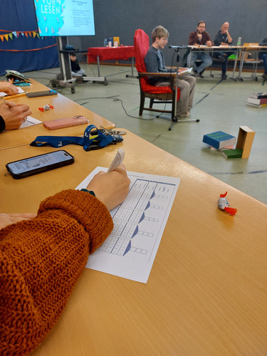 KI generiert: Das Bild zeigt einen Vorlesewettbewerb, bei dem ein Junge auf einem roten Stuhl sitzt und laut vorliest. Im Vordergrund notiert eine Person etwas auf einem Bewertungsformular, während im Hintergrund Jurymitglieder aufmerksam zuhören.