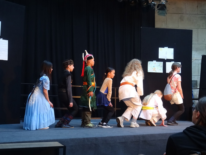 KI generiert: Das Bild zeigt eine Gruppe von Kindern in Kostümen auf einer Bühne, offenbar während einer Theateraufführung. Sie stehen in einer Linie und führen gemeinsam eine Szene auf.