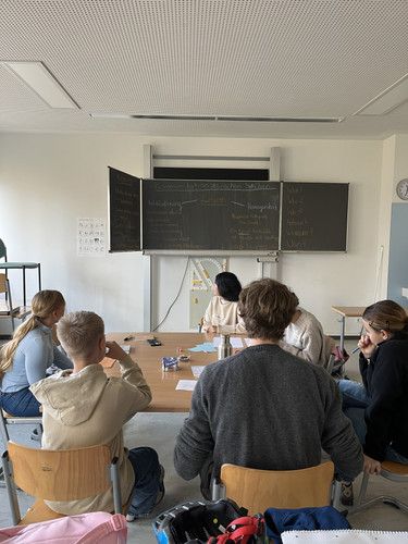 KI generiert: Das Bild zeigt eine Gruppe von Schülern, die in einem Klassenzimmer vor einer Tafel sitzen und wahrscheinlich an einer Diskussion oder Unterrichtsstunde teilnehmen. Auf der Tafel stehen Begriffe und Notizen, die auf das Thema Kommunikation schließen lassen.
