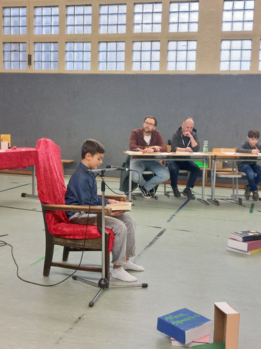 KI generiert: Ein Junge sitzt auf einem Stuhl und liest aus einem Buch vor, während er von drei Personen an einem Tisch beobachtet wird. Es scheint eine Vorleseveranstaltung oder ein Wettbewerb in einem Raum mit großen Fenstern zu sein.