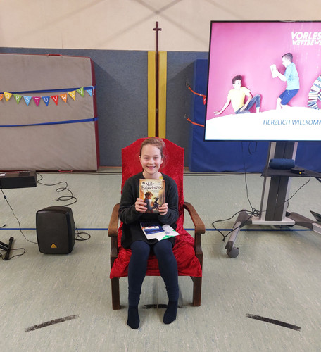 KI generiert: Auf dem Bild sitzt ein Kind auf einem roten Stuhl und hält ein Buch mit dem Titel "Nelly Rapp". Im Hintergrund sind eine Leinwand mit "Vorlesewettbewerb" und Dekorationen zu sehen.