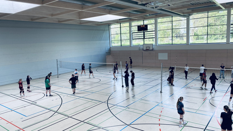 KI generiert: Das Bild zeigt eine Gruppe von Menschen, die in einer Sporthalle Volleyball spielen. Mehrere Teams sind auf zwei Spielfeldern aktiv und trainieren gemeinsam.