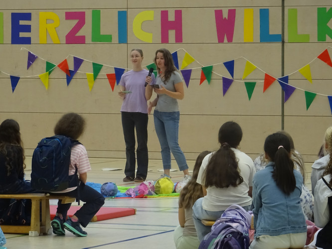 KI generiert: Das Bild zeigt zwei Personen, die offenbar eine Veranstaltung leiten und vor einer Gruppe von Kindern in einer Turnhalle sprechen. Im Hintergrund befinden sich bunte Dekorationen und der Schriftzug "Herzlich Willkommen".
