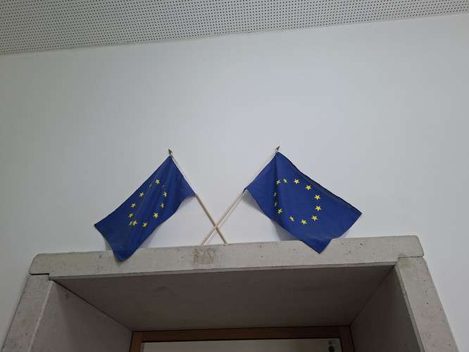 KI generiert: Das Bild zeigt zwei Europaflaggen, die überkreuz auf einem Betonelement an einer weißen Wand befestigt sind. Die Flaggen symbolisieren die Europäische Union und sind mit gelben Sternen auf blauem Hintergrund gestaltet.