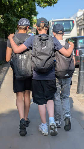 KI generiert: Drei Personen gehen auf einem Gehweg nebeneinander und legen freundschaftlich die Arme um die Schultern des jeweils anderen. Sie tragen Rucksäcke und Kappen.