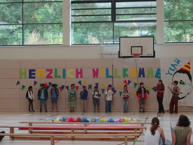 KI generiert: Das Bild zeigt eine Gruppe von Kindern, die in einer Turnhalle aufgereiht vor einem großen bunten "Herzlich Willkommen"-Banner stehen. Es scheint sich um eine Willkommensveranstaltung oder Einschulungsfeier zu handeln.