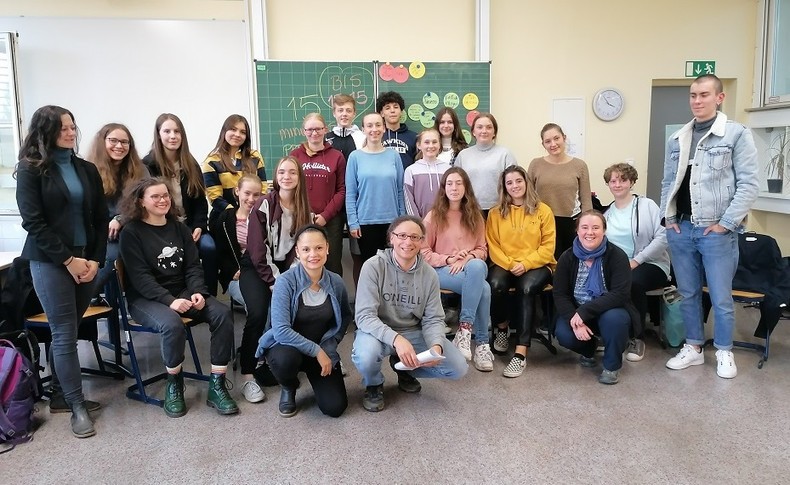 KI generiert: Eine Gruppe von Personen, hauptsächlich junge Erwachsene, posiert in einem Klassenraum vor einer Tafel für ein Gruppenfoto. Sie sind lächelnd und wirken entspannt in einer Lernumgebung.