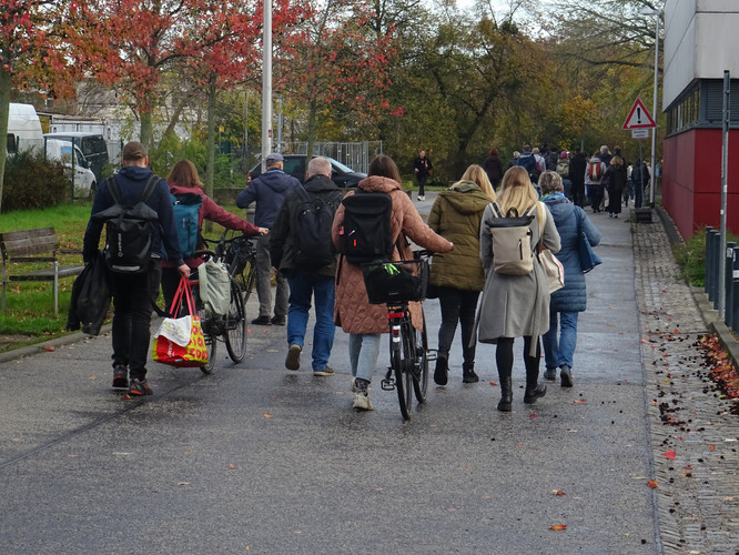 KI generiert: Eine Gruppe von Menschen, einige mit Fahrrädern, geht auf einem Gehweg entlang. Im Hintergrund sind Bäume mit herbstlichem Laub zu sehen.