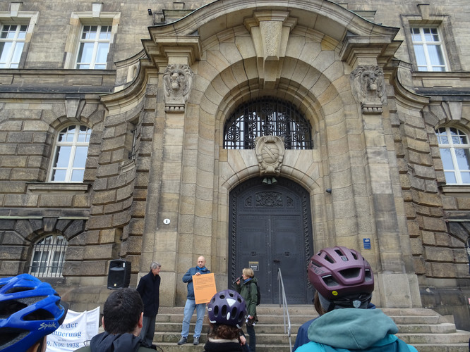 KI generiert: Das Bild zeigt eine Gruppe von Menschen, die vor einem alten, eindrucksvollen Gebäude mit großen Türen versammelt sind, wobei eine Person ein orangefarbenes Plakat hält. Einige der Anwesenden tragen Fahrradhelme.