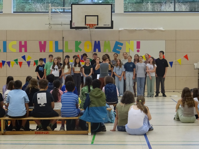 KI generiert: Das Bild zeigt eine Gruppe von Kindern oder Jugendlichen, die in einer Turnhalle vor Zuschauern stehen und möglicherweise eine Aufführung oder Präsentation machen. Im Hintergrund hängt ein "Herzlich Willkommen!"-Banner.