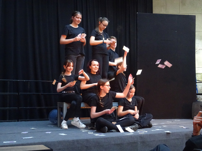 KI generiert: Auf dem Bild ist eine Gruppe von jungen Leuten in schwarzer Kleidung auf einer Bühne zu sehen, einige werfen mit Karten oder Zetteln in die Luft. Die Szene scheint Teil einer schauspielerischen oder künstlerischen Aufführung zu sein.