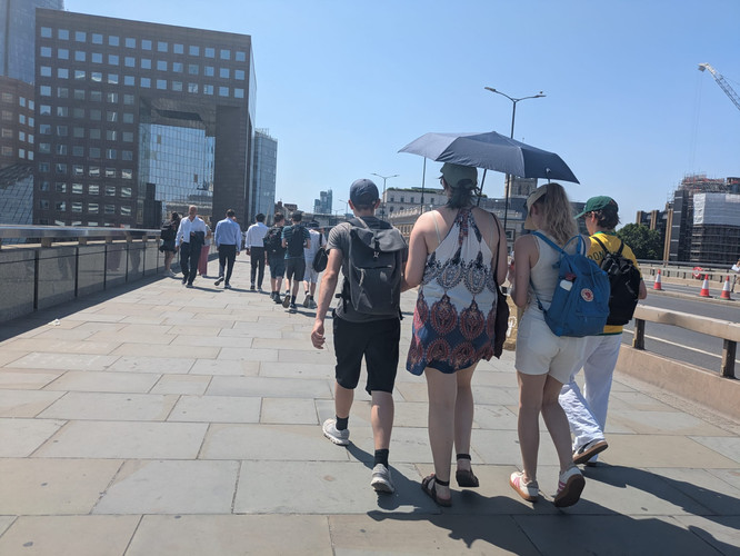 KI generiert: Das Bild zeigt eine Gruppe von Menschen, die auf einem Gehweg in einer urbanen Umgebung bei sonnigem Wetter gehen. Eine Frau hält einen Sonnenschirm, um sich vor der Sonne zu schützen.