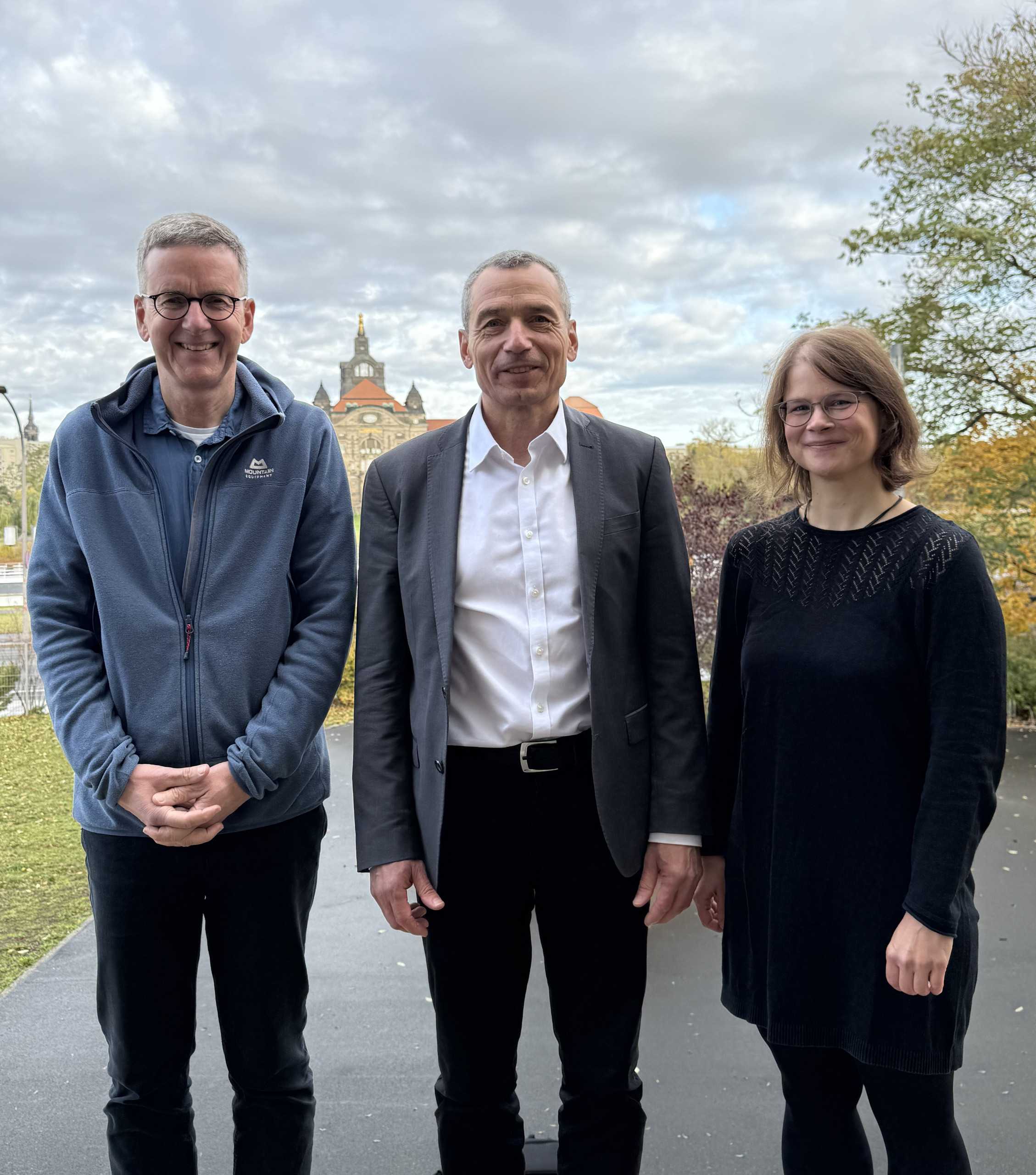 KI generiert: Das Bild zeigt drei Personen, die draußen nebeneinander stehen und in die Kamera lächeln. Im Hintergrund ist ein historisches Gebäude erkennbar, umgeben von herbstlicher Vegetation.