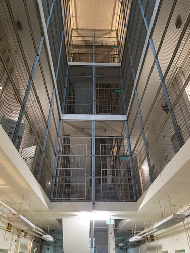 KI generiert: Das Bild zeigt das Innere eines Gefängnisses mit mehreren übereinander angeordneten Zellen, die durch Gitter getrennt sind. Der zentrale Flur führt zu höheren Ebenen des Gebäudes.