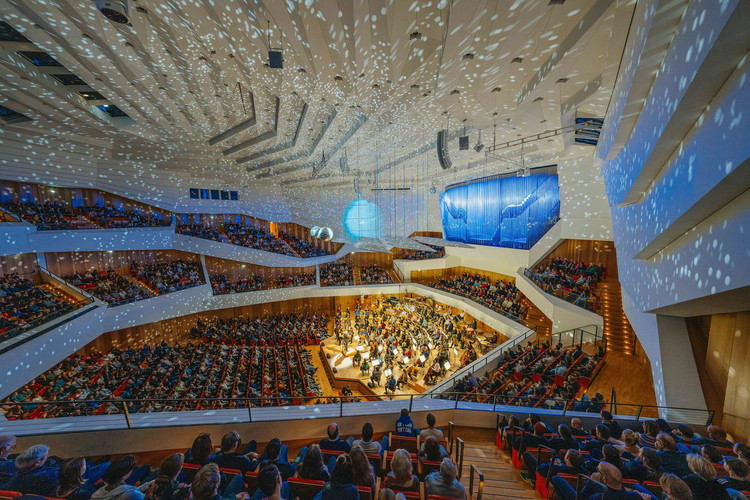 KI generiert: Das Bild zeigt einen großen Konzertsaal mit einem Orchester in der Mitte, umgeben von mehreren Ebenen an Sitzreihen, die gut gefüllt sind. Der Saal ist mit beeindruckenden Lichteffekten und einer großen Leinwand an der Wand ausgestattet, die dem Ambiente eine festliche Atmosphäre verleihen.