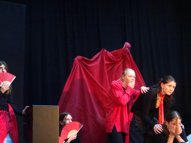 KI generiert: Das Bild zeigt eine Theateraufführung, bei der mehrere Personen in roten und schwarzen Kostümen posieren. Im Hintergrund hält eine Figur mit einem roten Umhang eine bedrohliche Position ein, während die anderen mit Gesichtsausdrücken der Überraschung oder Spannung reagieren.