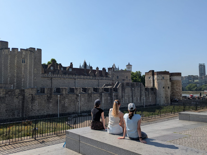 KI generiert: Drei Personen sitzen auf einer Mauer und blicken auf den Tower of London, eine historische Festungsanlage in einer urbanen Umgebung. Im Hintergrund sind die markanten Türme und Mauern des Gebäudes zu sehen.