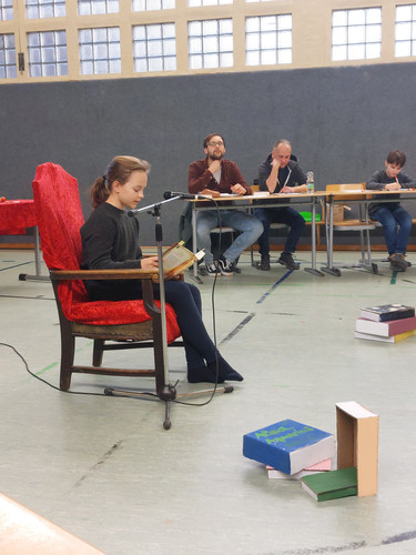 KI generiert: Ein Kind sitzt auf einem roten Stuhl und liest aus einem Buch vor einem Mikrofon vor, während einige Personen an Tischen im Hintergrund aufmerksam zuhören. Das Bild vermittelt den Eindruck einer Vorleseveranstaltung oder eines Wettbewerbs.