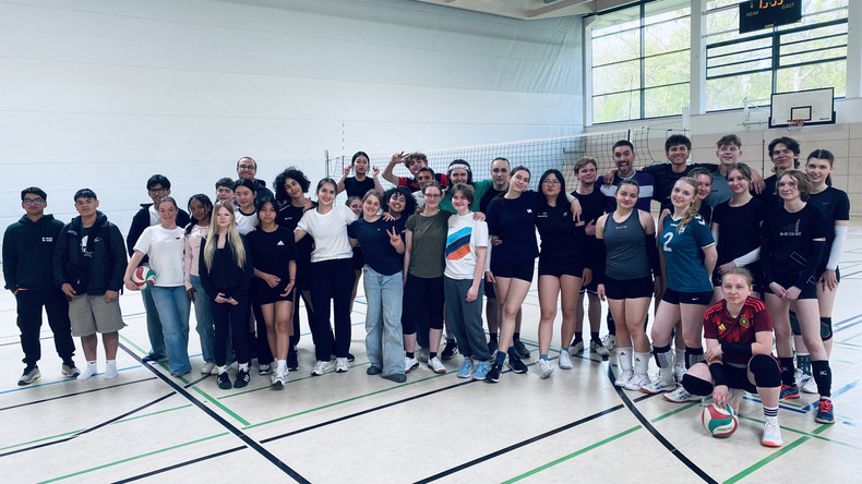 KI generiert: Das Bild zeigt eine große Gruppe von Menschen, die in einer Sporthalle posieren. Sie stehen teilweise in Sportkleidung um ein Volleyballnetz, was darauf hindeutet, dass es sich um ein Team- oder Klassenfoto im Rahmen einer Volleyballveranstaltung handelt.