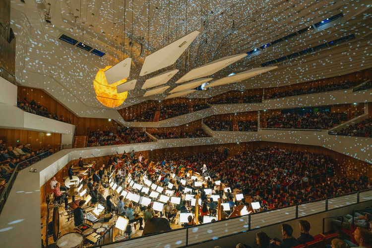 KI generiert: Das Bild zeigt ein voll besetztes Konzert in einem modernen Konzertsaal, beleuchtet von kreativen Lichteffekten. Ein großes Orchester spielt auf der Bühne unter einem beeindruckenden Kuppellicht.