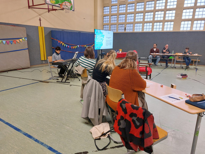 KI generiert: Das Bild zeigt eine Szene in einer Turnhalle, in der ein Mann auf einem Keyboard spielt und mehrere Personen an Tischen sitzen, anscheinend einem Vortrag oder einer Präsentation folgen. Im Hintergrund hängt ein Bildschirm mit dem Wort "Vorlesen".